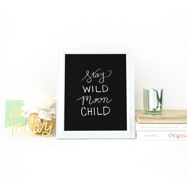 Stay Wild Moon Child Handlettered in white on black Print in a White Frame on a desk