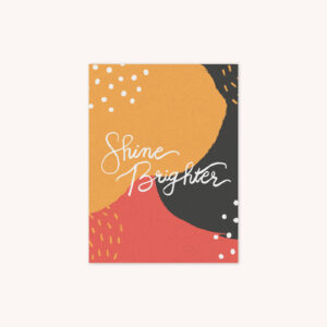 A greeting card featuring the phrase Shine Brighter handlettered in white on a yellow, red, and charcoal gray background with abstract shapes